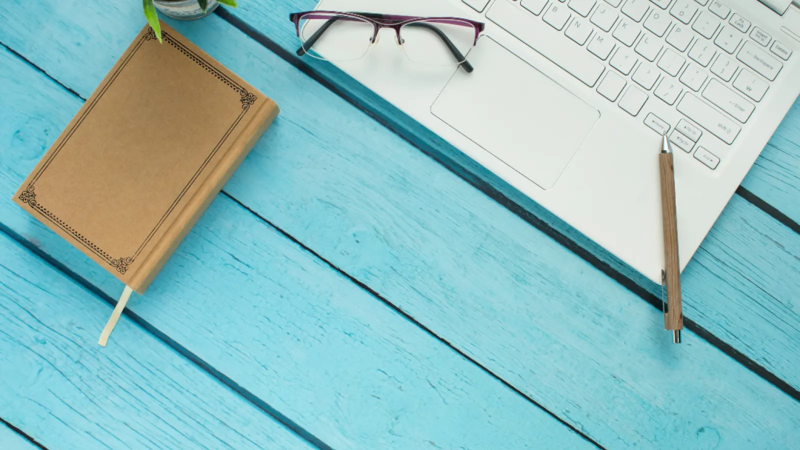 An Apple laptop sits on a blue table and a moleskin notebook lies nearby.